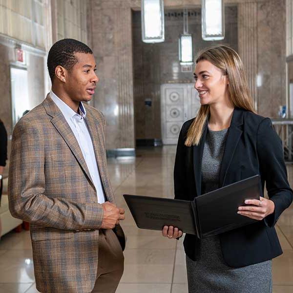 Two individuals standing in a hallway. On the left is a white woman with straight dark blonde hair. She wears a grey dress and a black jacket. She holds an open black folder. On the right is a Black man with short brown hair. He wears a tan-and-blue checkered suit and a white dress shirt